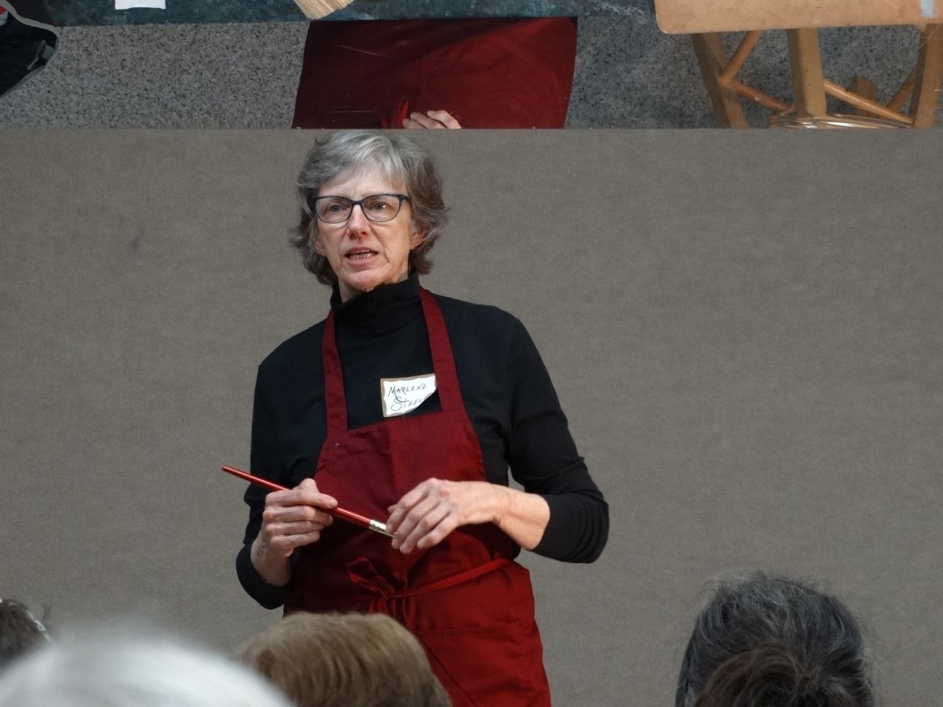 Marlene Steele speaking to an audience while holding a paintbrush